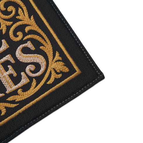 Rectangular black doormat with a low loop polyester pile and black edge trim framed by a thin gold rule, printed flat to imitate raised embroidery. Two almond shaped eyes with layered blue lids, cream whites, and blue irises ringed in gold gaze outward. Cream serif lettering with gold inlines reads THE EYE WATCHES. Gold scrollwork fills each corner, and a small gilded bird with outstretched wings sits at the bottom center.