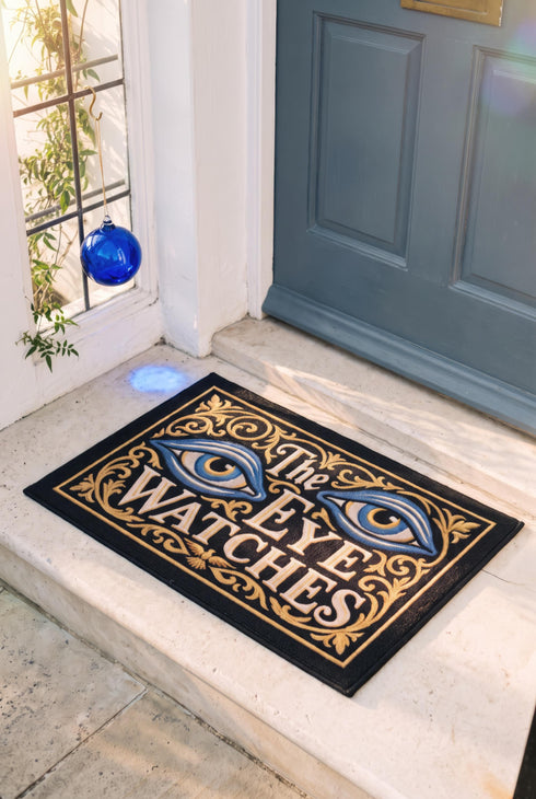 Rectangular black doormat with a low loop polyester pile and black edge trim framed by a thin gold rule, printed flat to imitate raised embroidery. Two almond shaped eyes with layered blue lids, cream whites, and blue irises ringed in gold gaze outward. Cream serif lettering with gold inlines reads THE EYE WATCHES. Gold scrollwork fills each corner, and a small gilded bird with outstretched wings sits at the bottom center.