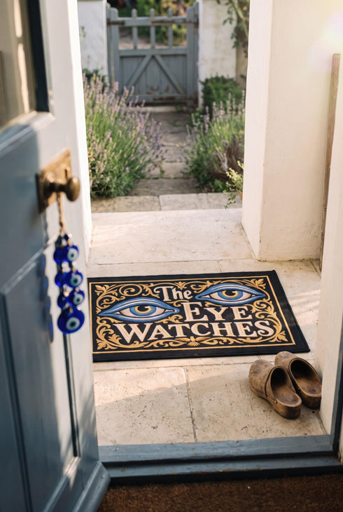 Rectangular black doormat with a low loop polyester pile and black edge trim framed by a thin gold rule, printed flat to imitate raised embroidery. Two almond shaped eyes with layered blue lids, cream whites, and blue irises ringed in gold gaze outward. Cream serif lettering with gold inlines reads THE EYE WATCHES. Gold scrollwork fills each corner, and a small gilded bird with outstretched wings sits at the bottom center.