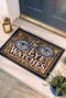 Rectangular black doormat with a low loop polyester pile and black edge trim framed by a thin gold rule, printed flat to imitate raised embroidery. Two almond shaped eyes with layered blue lids, cream whites, and blue irises ringed in gold gaze outward. Cream serif lettering with gold inlines reads THE EYE WATCHES. Gold scrollwork fills each corner, and a small gilded bird with outstretched wings sits at the bottom center.