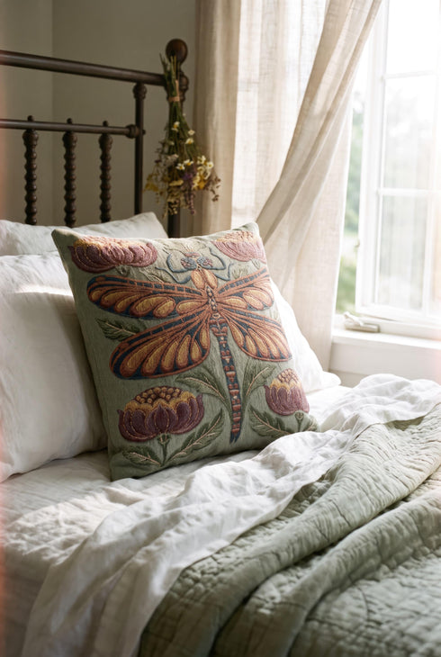 Square 17 by 18 inch jacquard woven throw pillow, woven identically on both faces and closed with a clean finished edge. A mirror symmetrical dragonfly fills the center of a sage green ground, its four long wings outlined in rust copper and filled with apricot and mustard, barred in slate blue along the leading edge. Plum peonies with ochre centers sit in each corner, and olive leaf sprays rise from the base. The ground has a soft sheen and the motif shows matte woven thread.