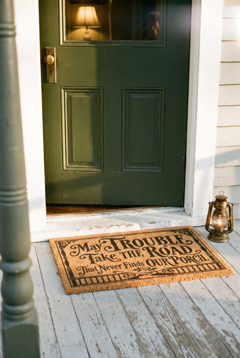 Rectangular natural tan coir doormat, 24 by 16 inches, with squared corners, a bristled tufted coconut fiber surface, and a black vinyl backing. The black artwork is printed into the fibers inside a double line frame. Ornate Victorian lettering reads MAY TROUBLE TAKE THE ROAD THAT NEVER FINDS OUR PORCH. Turned porch posts with scrolled corner brackets stand on each side, a row of balusters runs along the bottom, and a small winding road and magnolia blossom sit at the center.