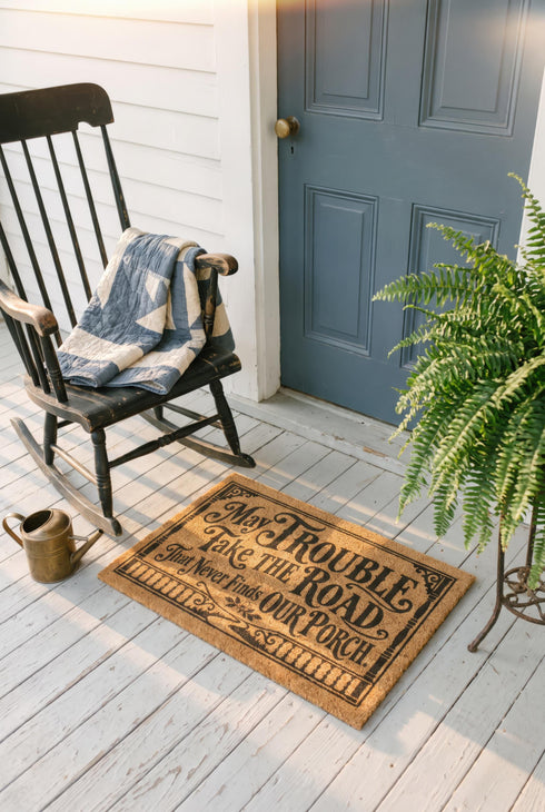 Rectangular natural tan coir doormat, 24 by 16 inches, with squared corners, a bristled tufted coconut fiber surface, and a black vinyl backing. The black artwork is printed into the fibers inside a double line frame. Ornate Victorian lettering reads MAY TROUBLE TAKE THE ROAD THAT NEVER FINDS OUR PORCH. Turned porch posts with scrolled corner brackets stand on each side, a row of balusters runs along the bottom, and a small winding road and magnolia blossom sit at the center.