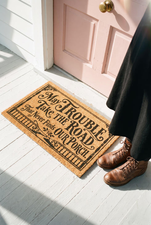 Rectangular natural tan coir doormat, 24 by 16 inches, with squared corners, a bristled tufted coconut fiber surface, and a black vinyl backing. The black artwork is printed into the fibers inside a double line frame. Ornate Victorian lettering reads MAY TROUBLE TAKE THE ROAD THAT NEVER FINDS OUR PORCH. Turned porch posts with scrolled corner brackets stand on each side, a row of balusters runs along the bottom, and a small winding road and magnolia blossom sit at the center.