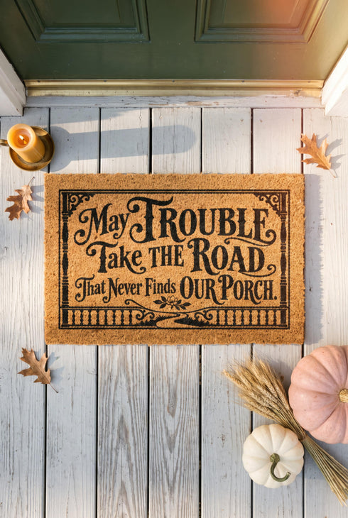 Rectangular natural tan coir doormat, 24 by 16 inches, with squared corners, a bristled tufted coconut fiber surface, and a black vinyl backing. The black artwork is printed into the fibers inside a double line frame. Ornate Victorian lettering reads MAY TROUBLE TAKE THE ROAD THAT NEVER FINDS OUR PORCH. Turned porch posts with scrolled corner brackets stand on each side, a row of balusters runs along the bottom, and a small winding road and magnolia blossom sit at the center.