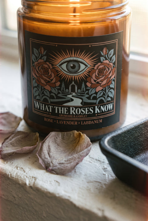 A 9 oz amber glass jar candle with a brushed gold screw lid and a wraparound black label. The label shows an all-seeing eye radiating peach and gold rays above a winding garden path that leads through slender cypress trees to an arched gate, flanked by two dusty coral roses on sage green stems, inside a fine copper border. Cream serif text reads What the Roses Know, Grimoire & Goblet, and Rose, Lavender, Labdanum. A romantic, witchy, art nouveau garden design.