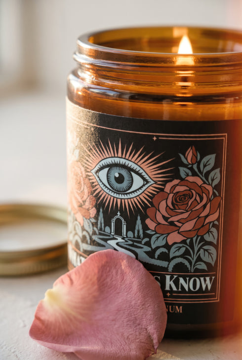 A 9 oz amber glass jar candle with a brushed gold screw lid and a wraparound black label. The label shows an all-seeing eye radiating peach and gold rays above a winding garden path that leads through slender cypress trees to an arched gate, flanked by two dusty coral roses on sage green stems, inside a fine copper border. Cream serif text reads What the Roses Know, Grimoire & Goblet, and Rose, Lavender, Labdanum. A romantic, witchy, art nouveau garden design.
