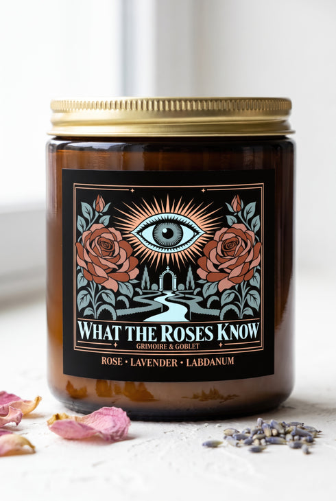 A 9 oz amber glass jar candle with a brushed gold screw lid and a wraparound black label. The label shows an all-seeing eye radiating peach and gold rays above a winding garden path that leads through slender cypress trees to an arched gate, flanked by two dusty coral roses on sage green stems, inside a fine copper border. Cream serif text reads What the Roses Know, Grimoire & Goblet, and Rose, Lavender, Labdanum. A romantic, witchy, art nouveau garden design.