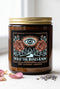 A 9 oz amber glass jar candle with a brushed gold screw lid and a wraparound black label. The label shows an all-seeing eye radiating peach and gold rays above a winding garden path that leads through slender cypress trees to an arched gate, flanked by two dusty coral roses on sage green stems, inside a fine copper border. Cream serif text reads What the Roses Know, Grimoire & Goblet, and Rose, Lavender, Labdanum. A romantic, witchy, art nouveau garden design.