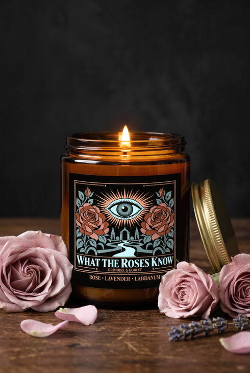 A 9 oz amber glass jar candle with a brushed gold screw lid and a wraparound black label. The label shows an all-seeing eye radiating peach and gold rays above a winding garden path that leads through slender cypress trees to an arched gate, flanked by two dusty coral roses on sage green stems, inside a fine copper border. Cream serif text reads What the Roses Know, Grimoire & Goblet, and Rose, Lavender, Labdanum. A romantic, witchy, art nouveau garden design.