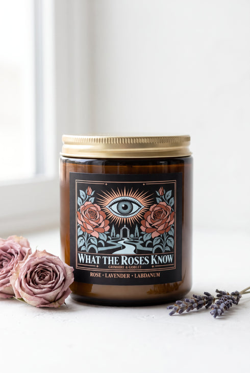 A 9 oz amber glass jar candle with a brushed gold screw lid and a wraparound black label. The label shows an all-seeing eye radiating peach and gold rays above a winding garden path that leads through slender cypress trees to an arched gate, flanked by two dusty coral roses on sage green stems, inside a fine copper border. Cream serif text reads What the Roses Know, Grimoire & Goblet, and Rose, Lavender, Labdanum. A romantic, witchy, art nouveau garden design.