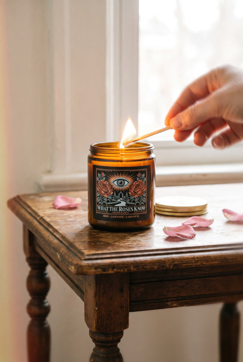 A 9 oz amber glass jar candle with a brushed gold screw lid and a wraparound black label. The label shows an all-seeing eye radiating peach and gold rays above a winding garden path that leads through slender cypress trees to an arched gate, flanked by two dusty coral roses on sage green stems, inside a fine copper border. Cream serif text reads What the Roses Know, Grimoire & Goblet, and Rose, Lavender, Labdanum. A romantic, witchy, art nouveau garden design.