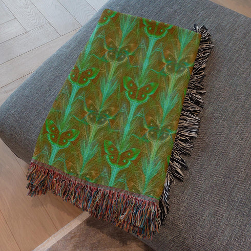 A woven cotton throw featuring an intricate luna moth design in deep green and russet tones, framed by soft fringe.